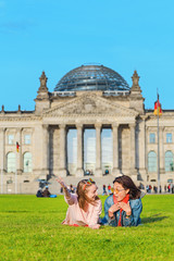 Fototapeta premium Dwie młode szczęśliwe dziewczyny w okularach przeciwsłonecznych leżące na trawie i bawią się przed budynkiem Bundestagu w Berlinie. Koncepcja studiów za granicą i podróży do Niemiec