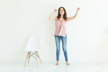 Emotions and people concept - young woman dancing near the white chair and smiling over the white background