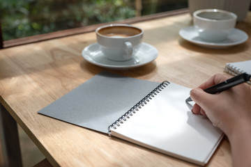 Closeup image of a hand writing down on a white blank notebook with coffee cup on wooden table