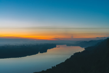 View from high shore on broad river. Riverbank with forest under thick fog. Dawn reflected in water. Yellow glow in picturesque predawn sky. Mystical morning atmospheric landscape of majestic nature.