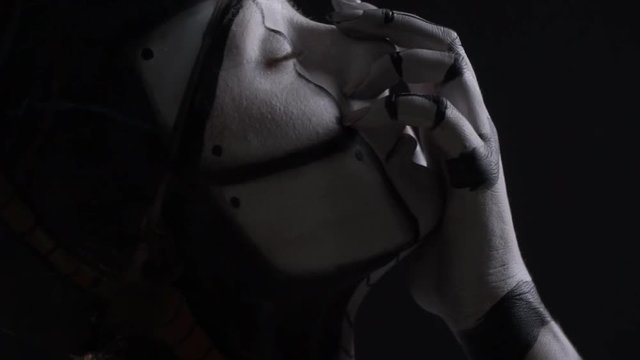 A cyborg woman stands with her eyes closed and touches her face