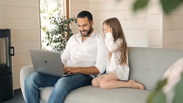 Young daughter sitting next to the father who working at the laptop, looking at the screen of device while man showing some images to the girl, indoor shot in modern apartment