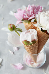 Decorative postcard of delicate pink and white peony flowers in a wafer cones in a glass vase with water drops, petals on a gray stone table.