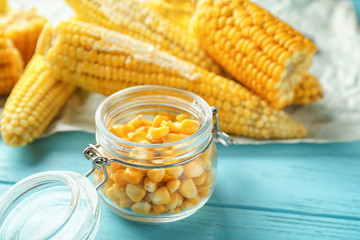 Glass jar with tasty corn kernels and cobs on wooden background, closeup