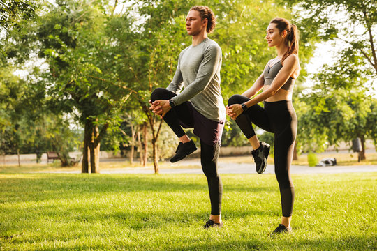 Photo of athletic man and woman 20s in tracksuits, warming up together and stretching legs in green park during sunny summer day