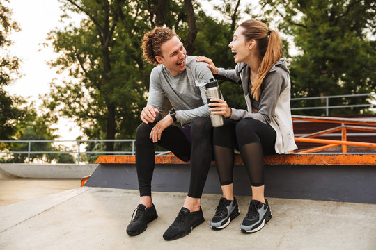 Image of happy sporty couple man and woman 20s in tracksuits, laughing while sitting on parapet in green park after training or fitness on sunny summer day
