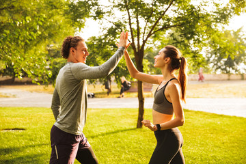 Photo of happy young man and woman 20s in tracksuits, doing workout together in green park during sunny summer day