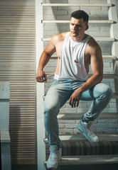 Obraz premium Guy in trendy tank top and jeans fashion. Young man sit on ladder. Athletic macho with muscular chest and hands. Fashion model with stylish hair. Mens fashion style and trend