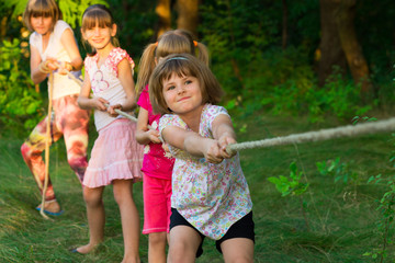 Group of happy children playing tug of war outside on grass. Kids pulling rope at park. Summer camp...
