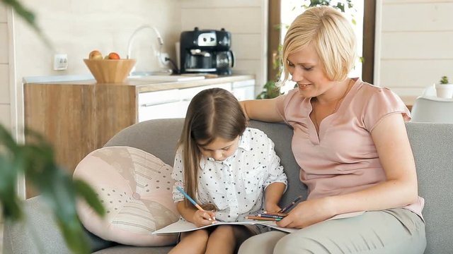 Caring mother drawing with daughter as sitting on nice gray coach, beautiful blond woman helping cute fair-haired kid with coloured pencils, indoor shot in studio apartment