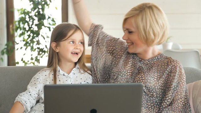 Caring mother watching cartoon with small daughter, enjoying spare time on comfortable gray sofa during summer holidays