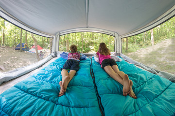 Bare footed children laying on blue sleeping bags in pop-up camper bunk end bed looking out window at surrounding forest trees and campground. Ultra wide angle 180 degree fisheye. 