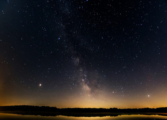 Milky Way galaxy in starry night sky over lake with reflections of trees in forest with light...