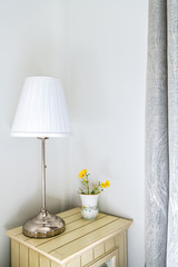 Closeup of modern nightstand in bedroom, room with side stainless steel lamp, flower vase, drapes, curtains in corner with walls