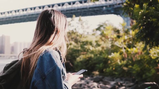 Side view of young traveler girl with backpack walking along calm Brooklyn Bridge river bank with smartphone slow motion