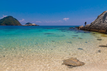 Fototapeta premium White fine sand submerged in glimmering shallow water, flanked by a big rock with a man on it & at the horizon Tukung Burung & the 2 mounds of the Susu Daras can be seen on Rawa beach, near Perhentian