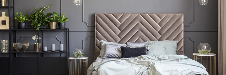 Bed with headboard between gold tables in grey bedroom interior with plants. Real photo - obrazy, fototapety, plakaty