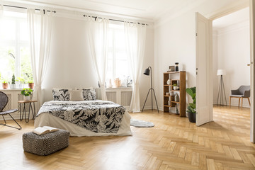 View at two rooms with white walls and herringbone parquet. A bed, a pouf, lamp bookcase and...