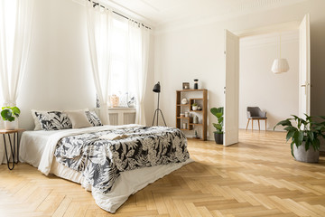 Stylish apartment interior with white walls and herringbone wooden floor. A view from a bedroom with a big bed to another room with an armchair. Real photo. - obrazy, fototapety, plakaty