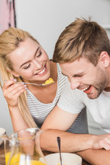 smiling girlfriend feeding laughing boyfriend with cornflakes in kitchen