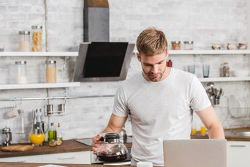 handsome man holding coffee pot and looking at laptop in kitchen