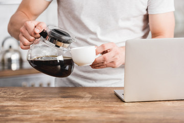 cropped image of man pouring coffee from coffee pot into cup near laptop in kitchen