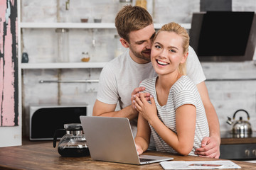 boyfriend hugging laughing girlfriend in kitchen while she working with laptop