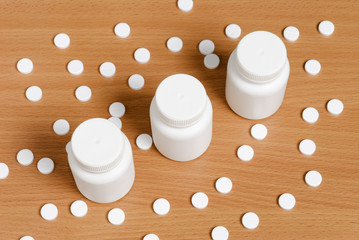 Fototapeta na wymiar White pills scattered on flat polished wooden surface around three bottles. Pills and bottles are white. Bottles are closed and arranged in row.