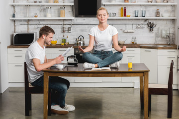 young man in earphones reading newspaper while beautiful girlfriend meditating on kitchen table