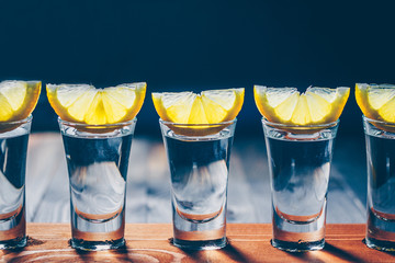 A row of wine glasses with vodka or tequila with lemon slices on a black background - obrazy, fototapety, plakaty