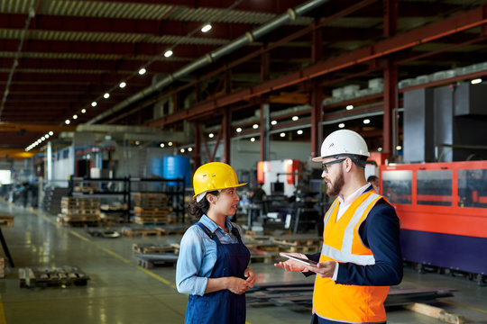 Side view portrait of production manager wearing hardhat talking to female factory worker standing in production workshop, copy space