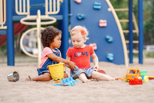 Give It To Me. Two Cute Caucasian And Hispanic Latin Babies Children Sitting In Sandbox Playing With Plastic Colorful Toys. Little Girls Friends Having Fun Together On Playground.