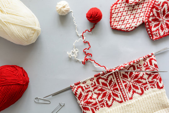 Girl knits red and white Norwegian jacquard hat knitting needles on gray wooden background. Process of knitting. Top view. Flat lay