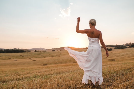 Young blond woman with white dress and short cut posing and walking by a dry straw countryside in a sunset of summer