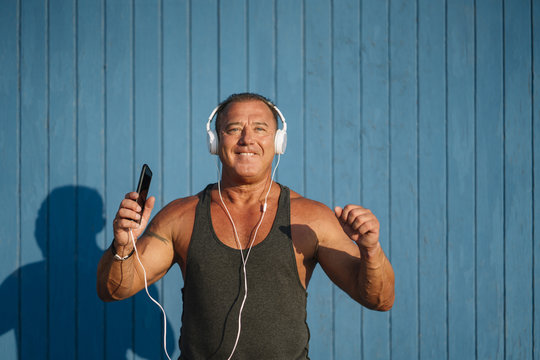 Strong older man poses with headphones on blue cabin background.