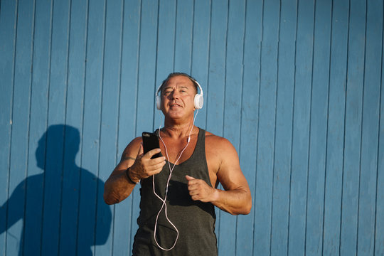 Strong older man poses with headphones on blue cabin background.