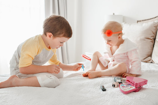 Adorable Caucasian boy and girl siblings playing together painting nails sitting on bed at home. Older brother doing manicure pedicure for his little younger sister. Friends toddlers using cosmetics.