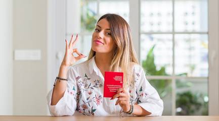 Young woman at home holding a passport of Switzerland doing ok sign with fingers, excellent symbol