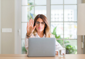 Young woman at home using laptop with open hand doing stop sign with serious and confident expression, defense gesture