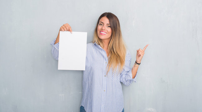 Young adult woman over grey grunge wall holding blank paper sheet very happy pointing with hand and finger to the side