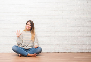 Young adult woman sitting on the floor over white brick wall at home showing and pointing up with fingers number five while smiling confident and happy.