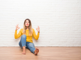 Young adult woman sitting on the floor over white brick wall relax and smiling with eyes closed doing meditation gesture with fingers. Yoga concept.