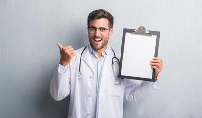 Handsome young doctor man over grey grunge wall holding clipboard pointing and showing with thumb up to the side with happy face smiling