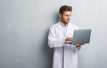 Handsome young man over grey grunge wall wearing white coat using laptop with a confident expression on smart face thinking serious