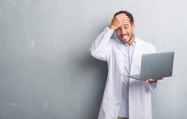 Handsome young man over grey grunge wall wearing white coat using laptop stressed with hand on...