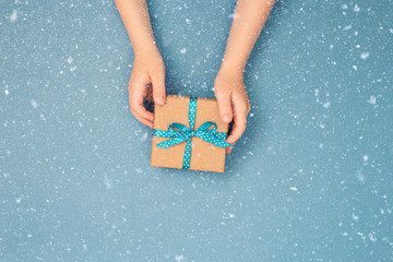 Child hands holding one gift box wrapped in kraft paper tied with blue ribbon in polka dots on blue grey background. Top view, place for text. Holiday concept.