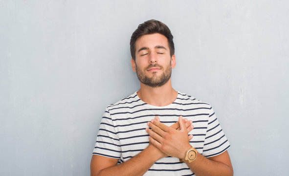 Handsome young man over grey grunge wall wearing navy t-shirt smiling with hands on chest with closed eyes and grateful gesture on face. Health concept.