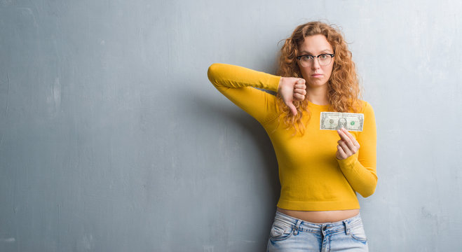 Young redhead woman over grey grunge wall holding a dollar with angry face, negative sign showing dislike with thumbs down, rejection concept