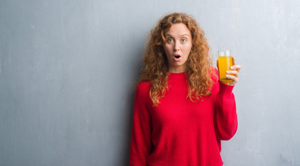 Young redhead woman over grey grunge wall drinking a glass of orange juice scared in shock with a...