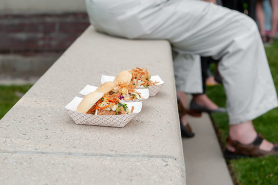 Three sandwiches in to-go cartons on a concrete ledge with fellow eaters in the background.